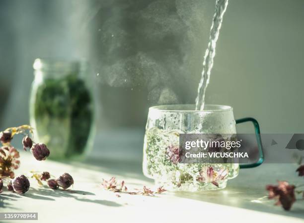 herbal tea with dried flowers in glass tea cup with fluent water and steam on table with petals and natural light - kräutertee stock-fotos und bilder