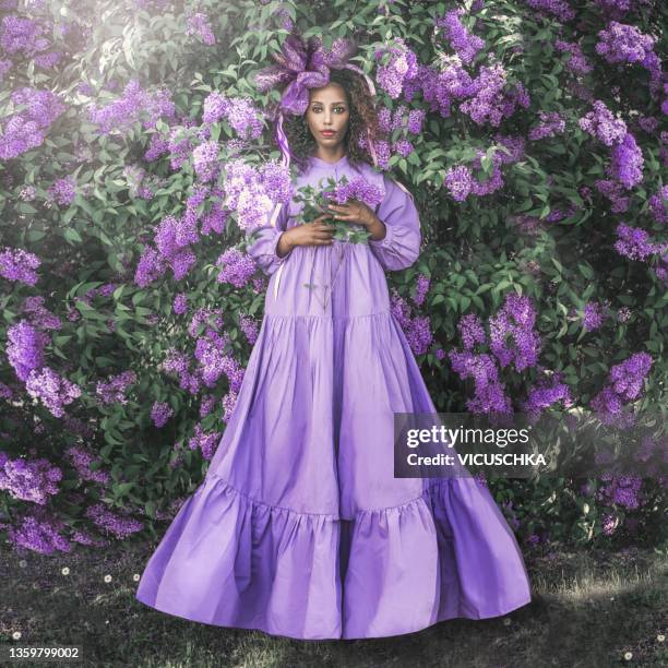 beautiful african young woman in long purple dress with flounces and headdress at lilac bushes with purple flowers bunch - purple lilac stock pictures, royalty-free photos & images