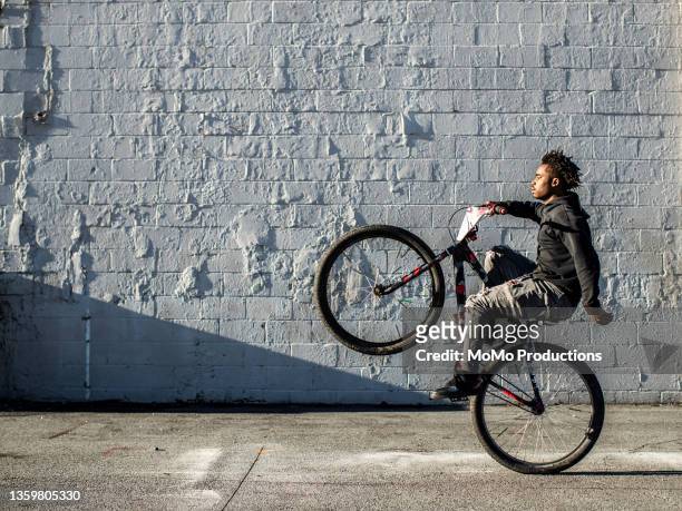young male bmx rider performing wheelie in urban area - wheelie stockfoto's en -beelden