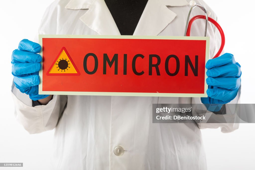 Doctor wearing latex gown and gloves holding a sign showing red coronavirus danger triangle and Omicron, on white background. Concept of coronavirus, health, pandemic, quarantine.
