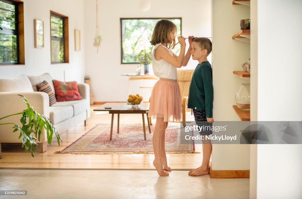 Girl measuring her younger brother's height against a wall at home