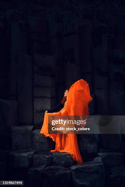 majestic scene of woman with red hair , wearing orange skirt flying it in the air on black rocky formations - orange skirt stock pictures, royalty-free photos & images