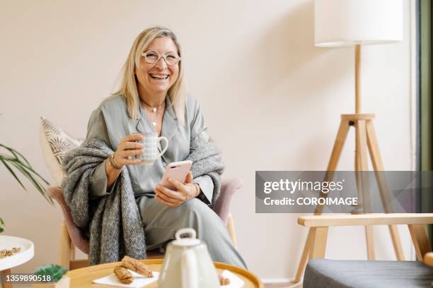 joyful and relaxed woman using her mobile phone while drinking a cup of coffee at home. - period cup stockfoto's en -beelden