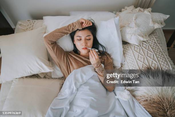 directly above asian chinese woman lying on bed covered with blanket having  cold and flu measured her body temperature with digital thermometer - illness stockfoto's en -beelden