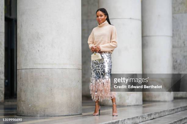 Ellie Delphine wears gold large earrings, a beige ribbed wool turtleneck oversized pullover, a silver large sequins high waist tube midi skirt with...