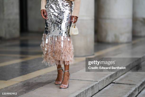 Ellie Delphine wears a beige ribbed wool oversized pullover, a silver large sequins high waist tube midi skirt with pale pink feathers on the ankle,...