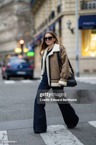 Diane Batoukina wears black sunglasses, a black t-shirt, a brown leather with black strap and white sheep interior oversized aviator coat, high waist...