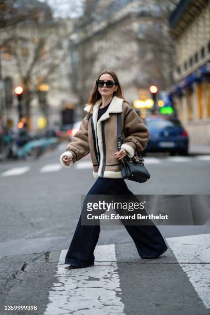 Diane Batoukina wears black sunglasses, a black t-shirt, a brown leather with black strap and white sheep interior oversized aviator coat, high waist...