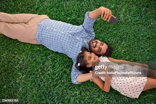 a father and daughter clicking selfie on mobile while relaxing on grass - father clicking selfie bildbanksfoton och bilder