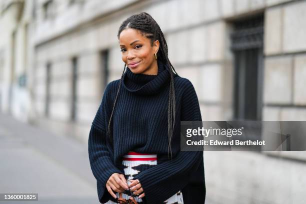 Ellie Delphine wears a black braided ribbed wool turtleneck oversized pullover, a high waits white with black and gold print pattern and purple blue...