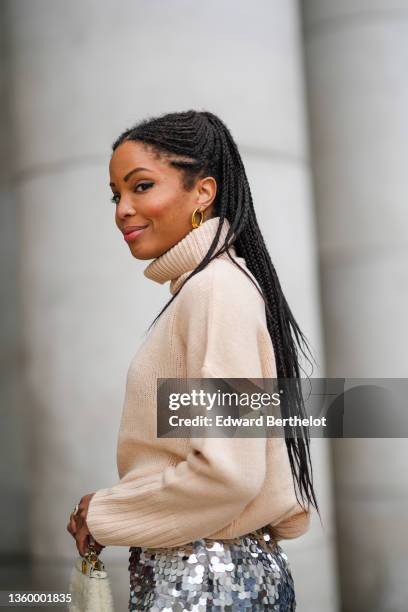 Ellie Delphine wears gold large earrings, a beige ribbed wool turtleneck oversized pullover, a silver large sequins high waist tube midi skirt with...