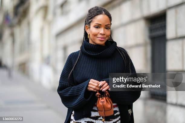 Ellie Delphine wears a black braided ribbed wool turtleneck oversized pullover, gold earrings, a high waits white with black and gold print pattern...