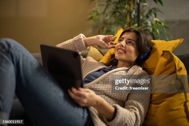 mujer joven acostada en el sofá y escuchando música en auriculares - mujeres de mediana edad fotografías e imágenes de stock