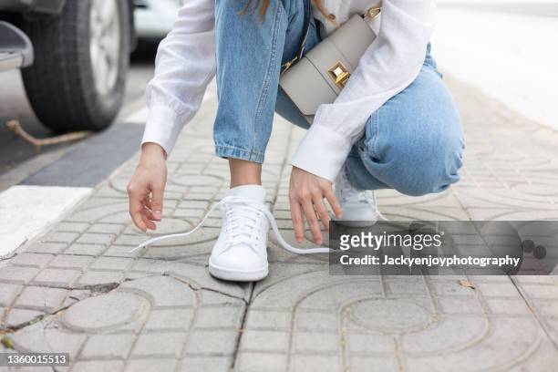 the girl's feet are in new white sneakers and jeans. a woman in athletic shoes squats down and ties an untied shoelace. a fashionable and stylish lifestyle. - amarrar o cadarço - fotografias e filmes do acervo