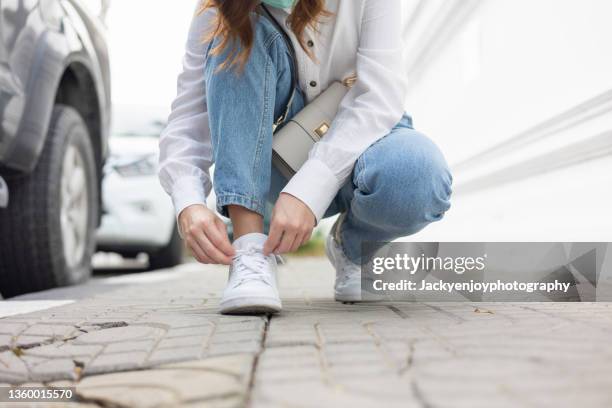 the girl's feet are in new white sneakers and jeans. a woman in athletic shoes squats down and ties an untied shoelace. a fashionable and stylish lifestyle. - untied shoelace stock pictures, royalty-free photos & images