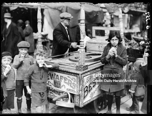Children with ice cream seller, about 1936. A photograph of a group of children gathered around an ice cream seller and enjoying ice cream wafers,...