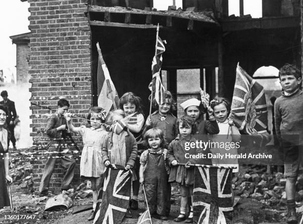 Children waving flags on VE Day, 8 May 1945. 'In bomb scarred Battersea, the little Londoners celebrated V E Day among the ruins of their homes'.