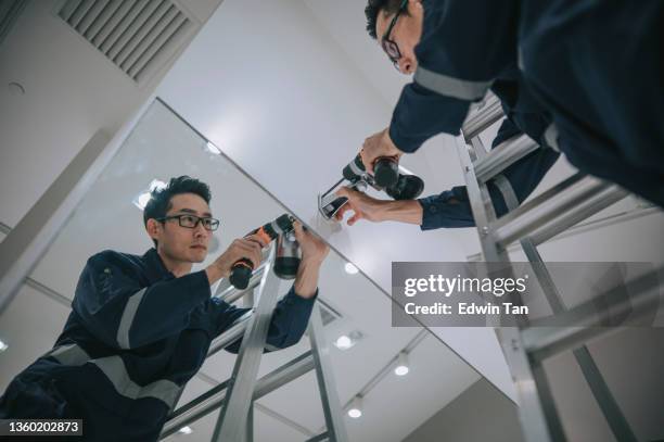 un homme asiatique chinois de milieu adulte grimpe sur une échelle installant une caméra de sécurité cctv dans un magasin de vêtements derrière le système de point de vente de comptoir sur montage mural - camera de surveillance photos et images de collection