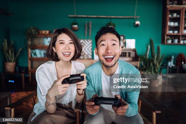 feliz joven pareja asiática sentada en el sofá de la sala de estar, divirtiéndose jugando videojuegos juntos en casa - partido de fútbol fotografías e imágenes de stock