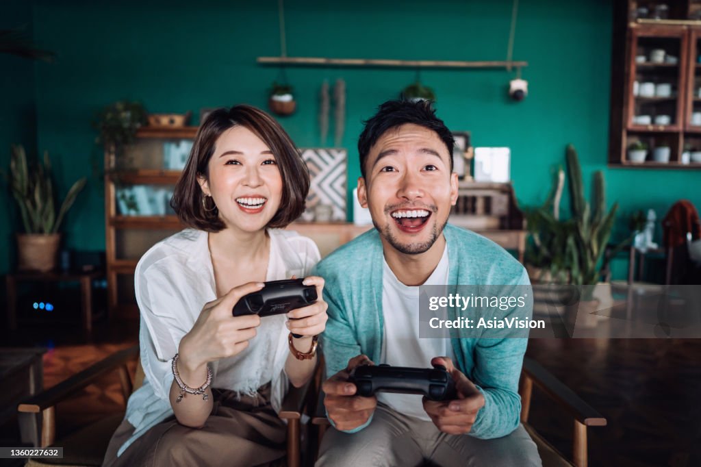 Happy young Asian couple sitting on the sofa in the living room, having fun playing video games together at home