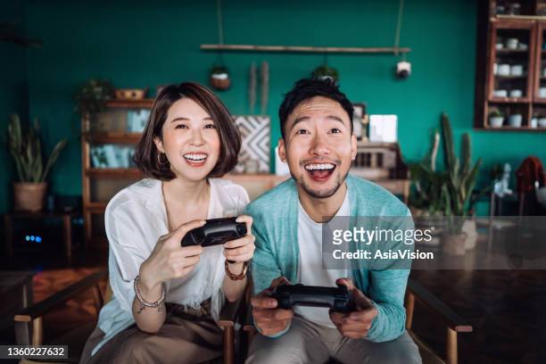 happy young asian couple sitting on the sofa in the living room, having fun playing video games together at home - far east stock pictures, royalty-free photos & images