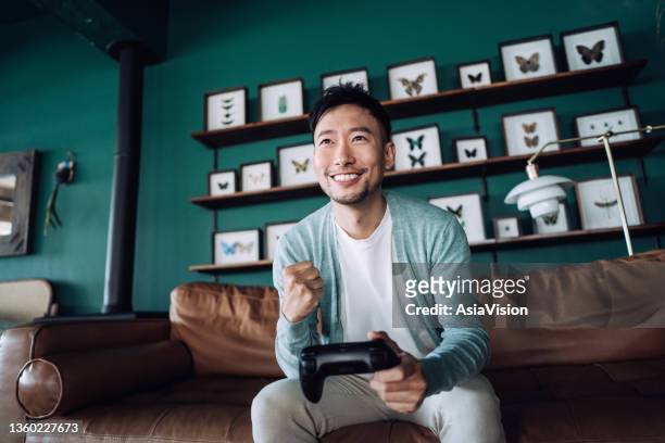 happy young asian man sitting on the sofa in the living room, shouting in excitement while having fun playing video games at home - sports man cave stock pictures, royalty-free photos & images