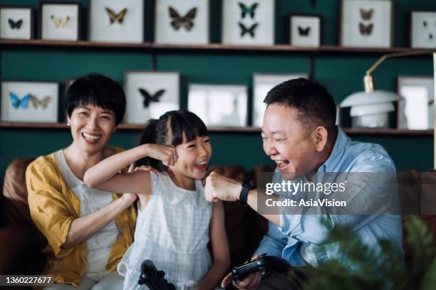 cheerful asian grandparents and granddaughter sitting on the sofa in the living room, shouting in excitement and giving each other a fist pump while having fun playing video games together at home. happy retirement concept - child fist bump stock pictures, royalty-free photos & images