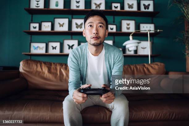konzentrierter junger asiatischer mann, der auf dem sofa im wohnzimmer sitzt und spaß daran hat, zu hause videospiele zu spielen - man cave stock-fotos und bilder