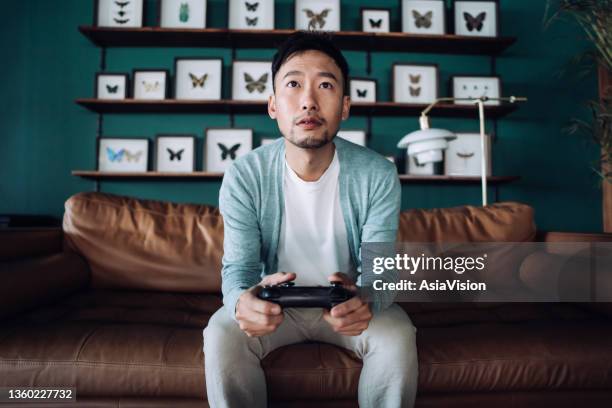 concentrated young asian man sitting on the sofa in the living room, having fun playing video games at home - chinese fan stock pictures, royalty-free photos & images