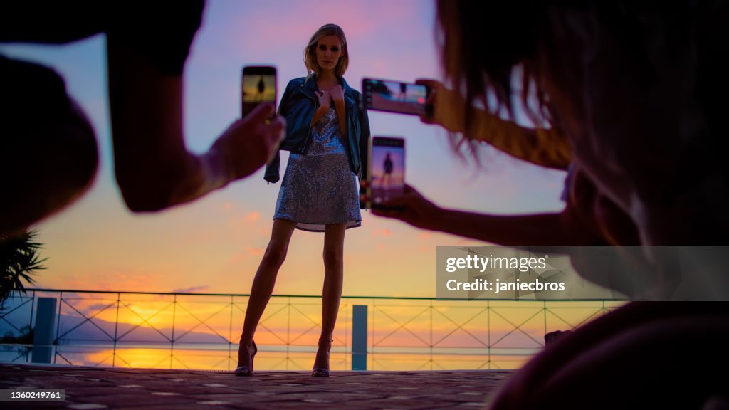 Romantischer Sonnenuntergang auf einer Terrasse. Schöne Frau posiert im Paillettenkleid für Fotos von Smartphones