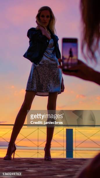 romantischer sonnenuntergang auf einer terrasse. schöne frau posiert im paillettenkleid für smartphone-foto - paillettenkleid stock-fotos und bilder