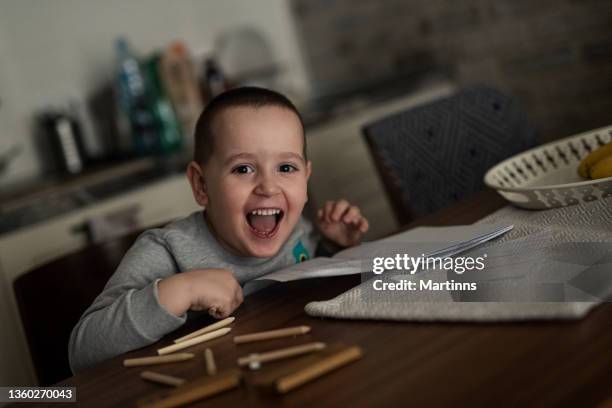 smiling young boy sitting at the kitchen table, drawing on pa paper and having fun - bruine ogen stockfoto's en -beelden