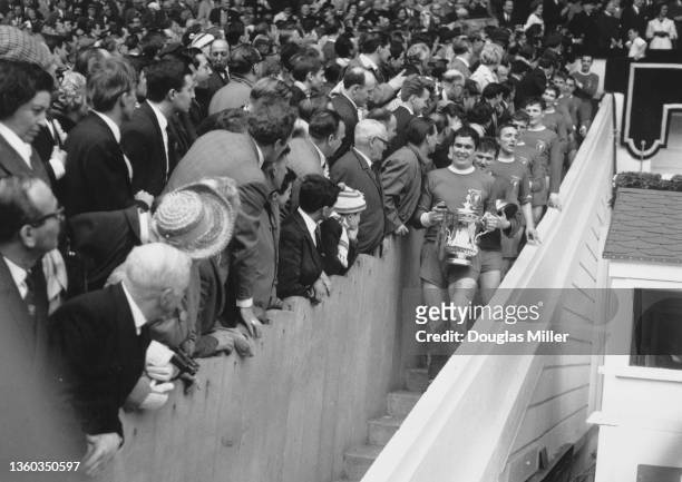 Fans look on as Liverpool Football Club team captain Ron Yeats walks down the steps from the royal box holding the Football Association trophy...