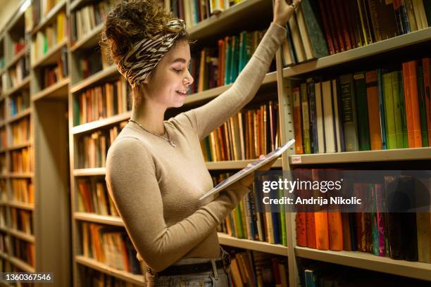 junge studentin sucht in der bibliothek in der hand nach einem buch und hält tablett - literatur stock-fotos und bilder