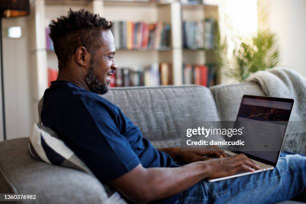 relaxed smiling man sitting on sofa and using laptop - subscription stockfoto's en -beelden