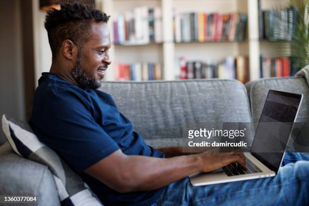 hombre sonriente relajado sentado en el sofá y usando la computadora portátil - web fotografías e imágenes de stock