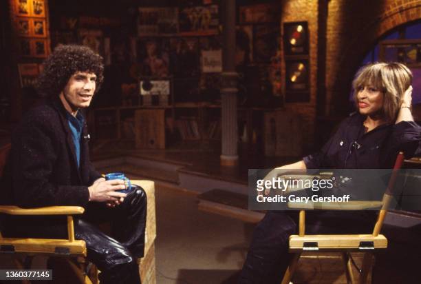 View of American VJ Mark Goodman and R&B, Rock, and Pop singer Tina Turner as they sit in director's chairs during an interview on MTV at Teletronics...