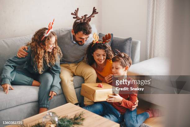 eine junge familie sitzt im wohnzimmer. eltern und ihre kinder haben ein geweih auf dem kopf. ein kleiner junge hält ein weihnachtsgeschenk in der hand. - antler stock-fotos und bilder