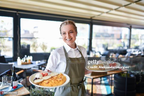 beautiful smiling blond waitress bringing two plates of scrambled eggs breakfast to restaurant table - ober stockfoto's en -beelden