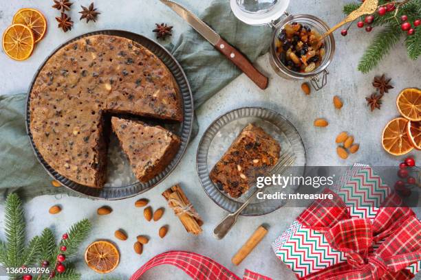 image of sliced, homemade, christmas fruitcake on plate, jar of mincemeat, wrapped gift with bow, knife, green muslin, cinnamon stick bundles, almonds, dried citrus orange fruit, red berries, spruce needles, star anise, elevated view - christmas cake stock pictures, royalty-free photos & images