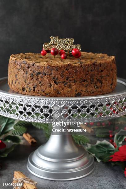 image of homemade, christmas fruitcake decorated with gold merry christmas topper and red berries, on silver cake stand surrounded by artificial poinsettias and variegated holly, spruce needles and red berries, cinnamon stick bundle, focus on foreground - christmas cake stock pictures, royalty-free photos & images
