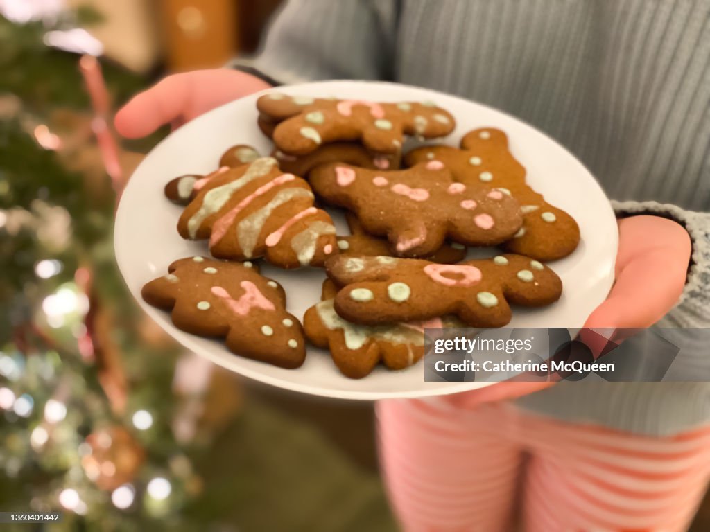 Mixed-race female holds plate of gingerbread cookies with illuminated christmas tree in background