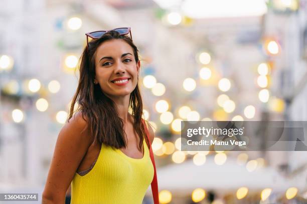 confident positive young brunette with long hair wearing yellow dress and trendy sunglasses over the head looking at camera while standing against blurred urban background with sparkling lights - portrait blurred background stockfoto's en -beelden