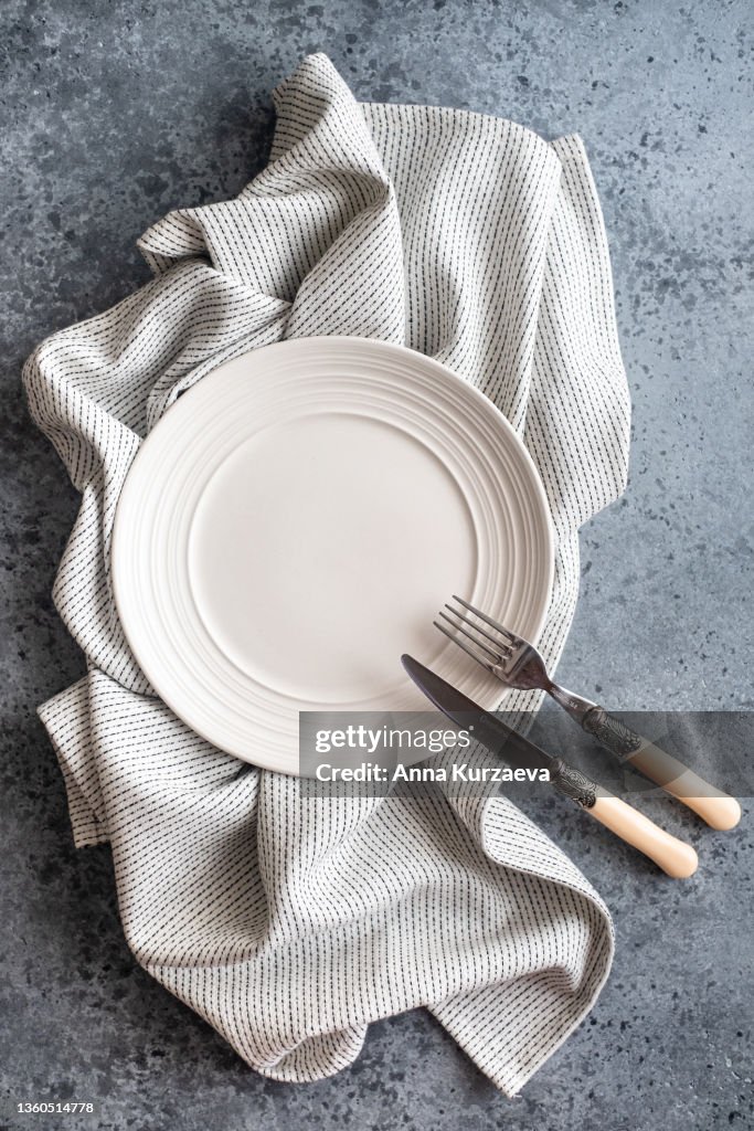 Empty white plate with fork and knife on a white and black linen striped napkin folded on grey concrete background, top view. Copy space