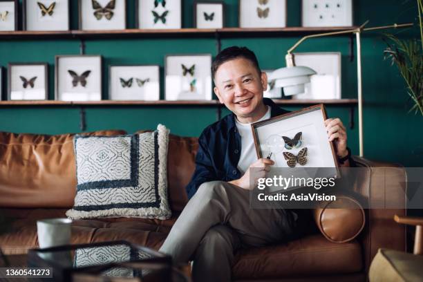 portrait of cheerful senior asian man relaxing on sofa at cozy home, holding insect collection specimen holder with magnifying glass, looking at camera with smile. happy retirement life, hobbies and interests for the elderly concept - collections stock pictures, royalty-free photos & images