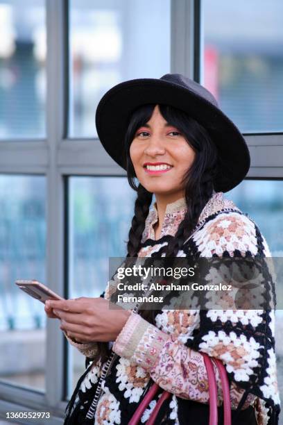 vertical image of an ecuadorian woman with braids, black hat and a traditional poncho looking at camera while using her mobile phone in the city - ecuadorian ethnicity stock pictures, royalty-free photos & images