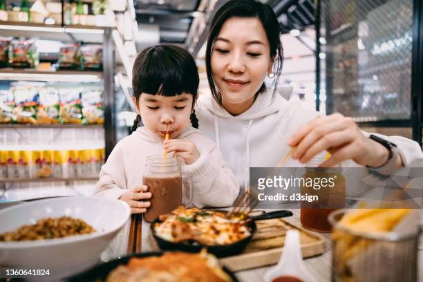 Young Asian mother and lovely little daughter enjoying meal together in restaurant, with assorted scrumptious food freshly served on the dining table. Family and eating out lifestyle