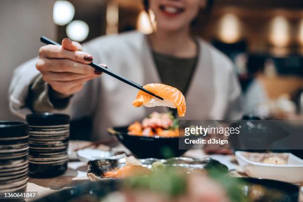 close up, mid-section of smiling young asian woman enjoying delicate freshly served sushi with chopsticks in japanese restaurant, sharing assorted authentic japanese cuisine with friends while dining out. asian cuisine and food. people and food concept - woman sushi stock-fotos und bilder