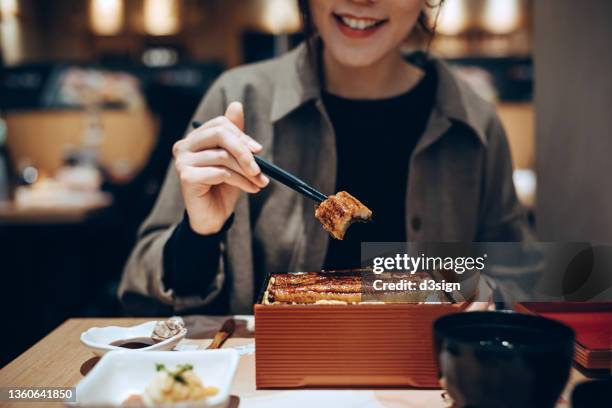 close up, mid-section of smiling young asian woman enjoying traditional grilled unagi over rice in box in japanese restaurant. authentic japanese cuisine and food culture. people and food concept - bento stock pictures, royalty-free photos & images