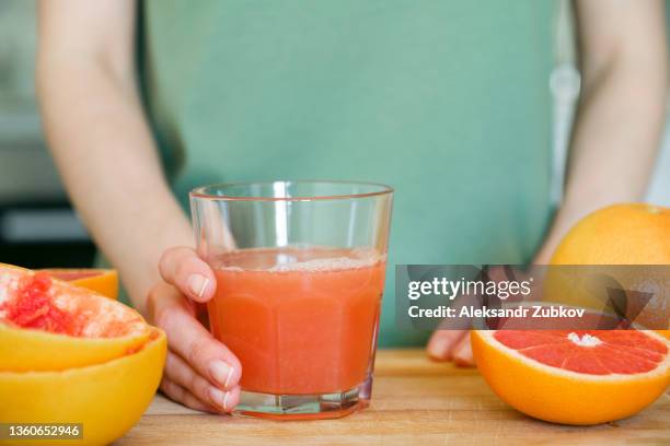 a girl or a woman is holding a glass of freshly squeezed juice from a red ripe grapefruit, on a cutting board, against the background of a wooden kitchen table. i make juice for breakfast. the concept of vegetarian, vegan and raw food. - immune system stock pictures, royalty-free photos & images